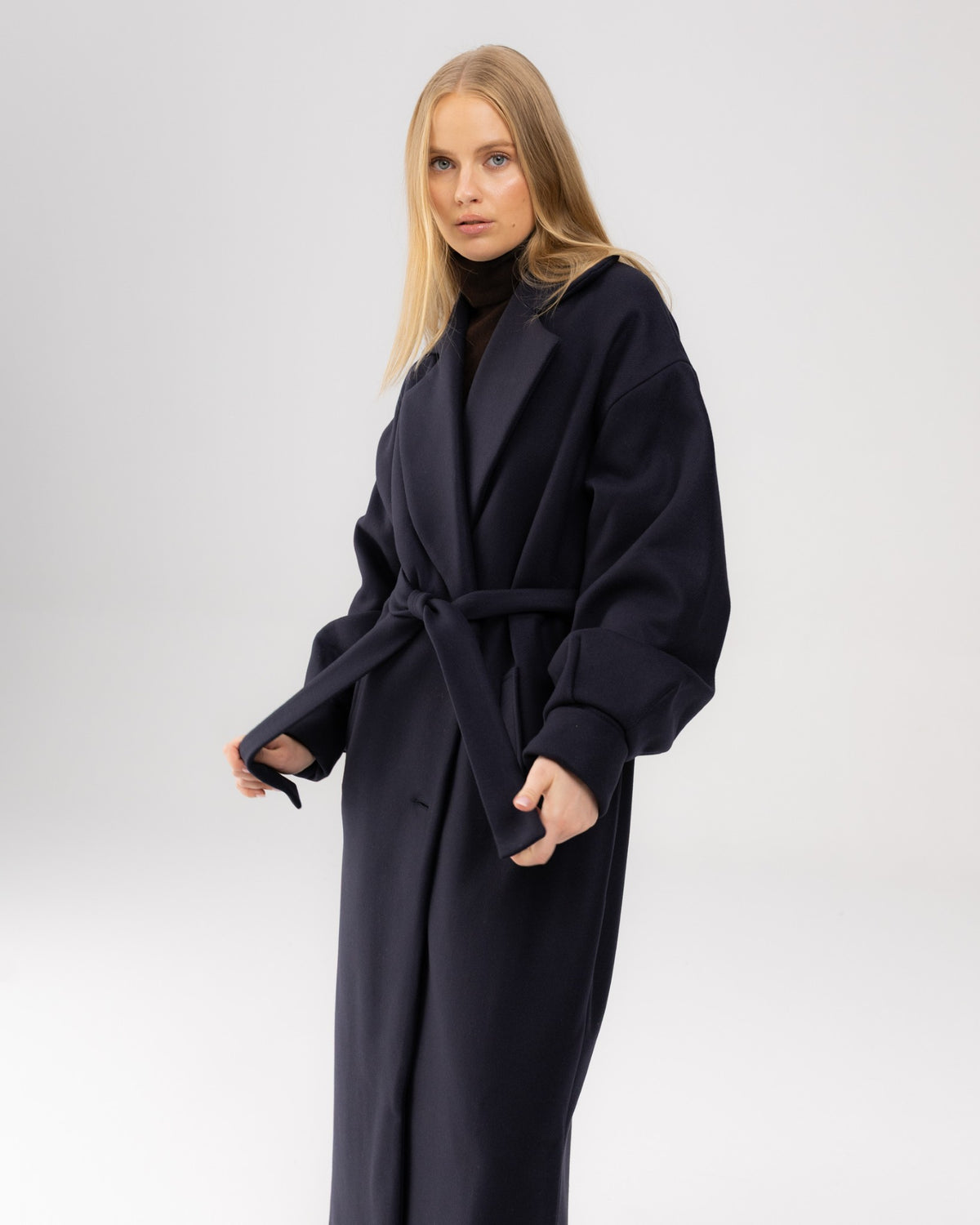 Granatowy wełniany płaszcz oversize na modelce trzymającej pasek, ujęcie do kolan